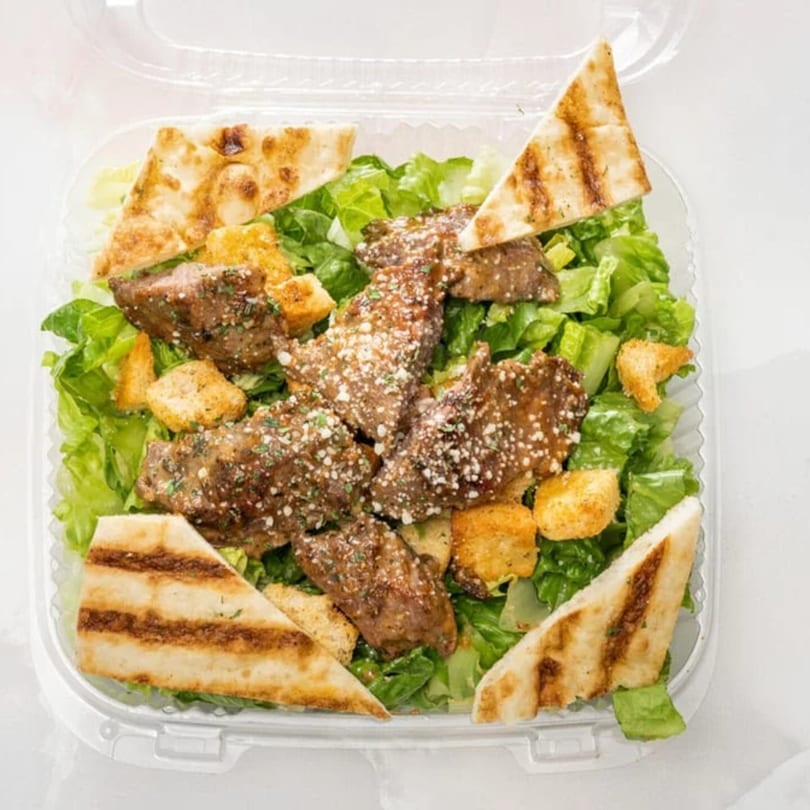 Steak Caesar salad with grilled pita bread and croutons in a clear plastic container.