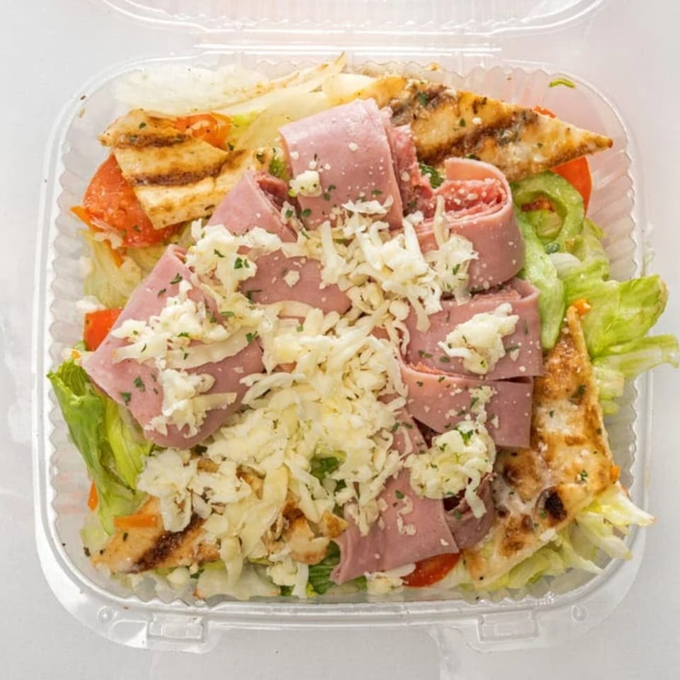 A clear plastic container filled with a fresh salad, topped with ham, grilled chicken, and shredded cheese.