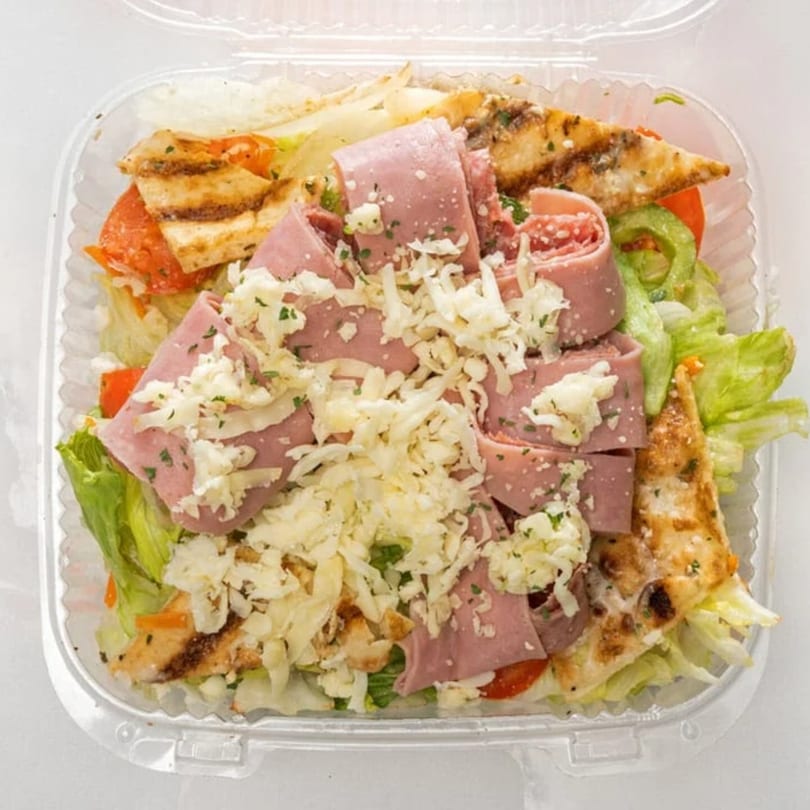 A clear plastic container filled with a fresh salad, topped with ham, grilled chicken, and shredded cheese.